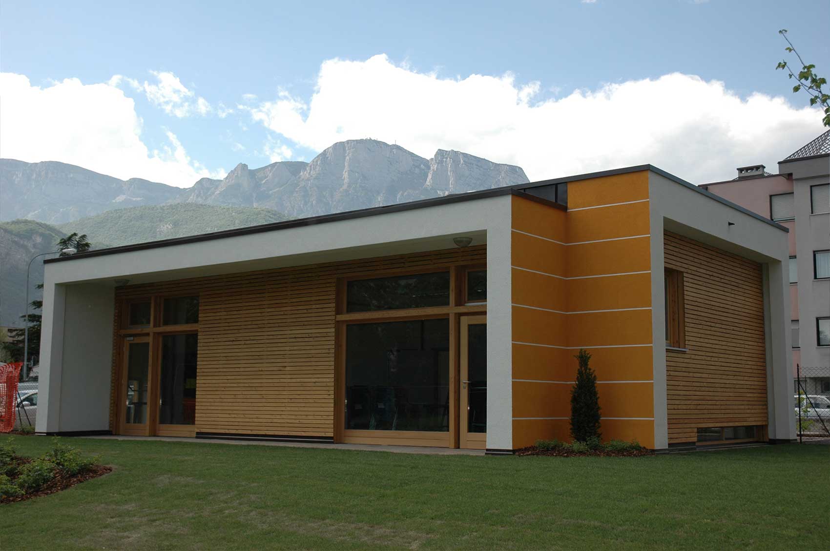 Haus Gardasee mit Flachdach, Holzfassade und orangefarbenem Fassadenelement vor einer Berglandschaft